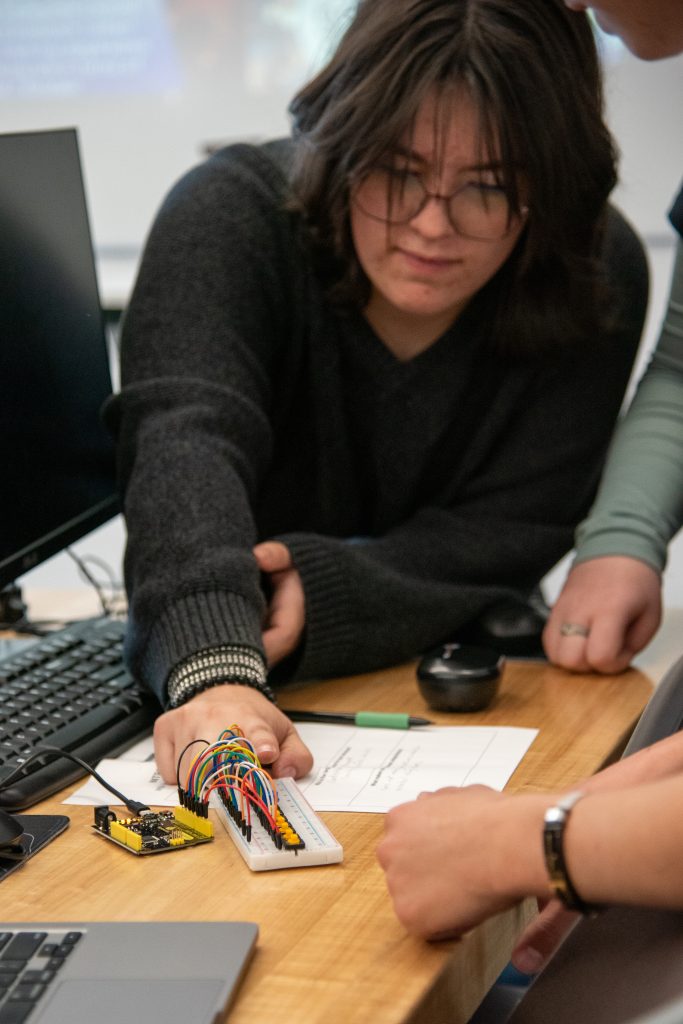 A student works on a circuit with wires on a breadboard, with a laptop and notes visible on the table.