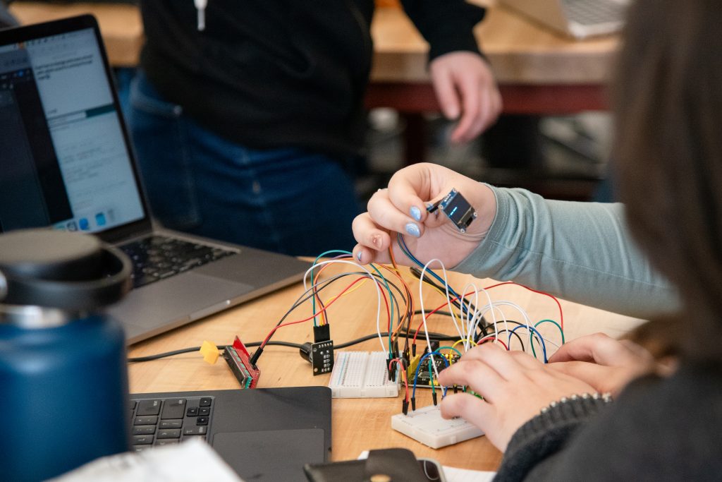 A hand holds a small display while another hand connects wires to a circuit board, with a laptop nearby.