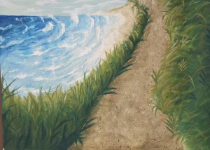 A sandy path lined with green grass leads to a blue ocean under a partly cloudy sky.