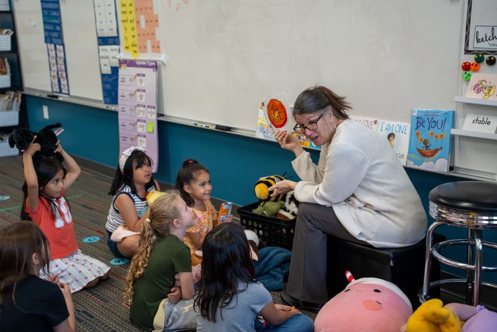 A teacher engages a small group of children with a story in a colorful classroom filled with books and toys.