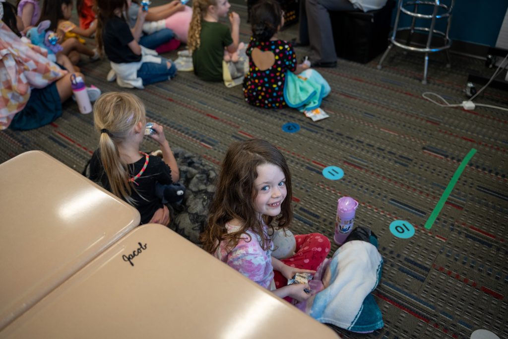 A young girl with curly hair smiles, sitting on the floor among other children, during a group activity.