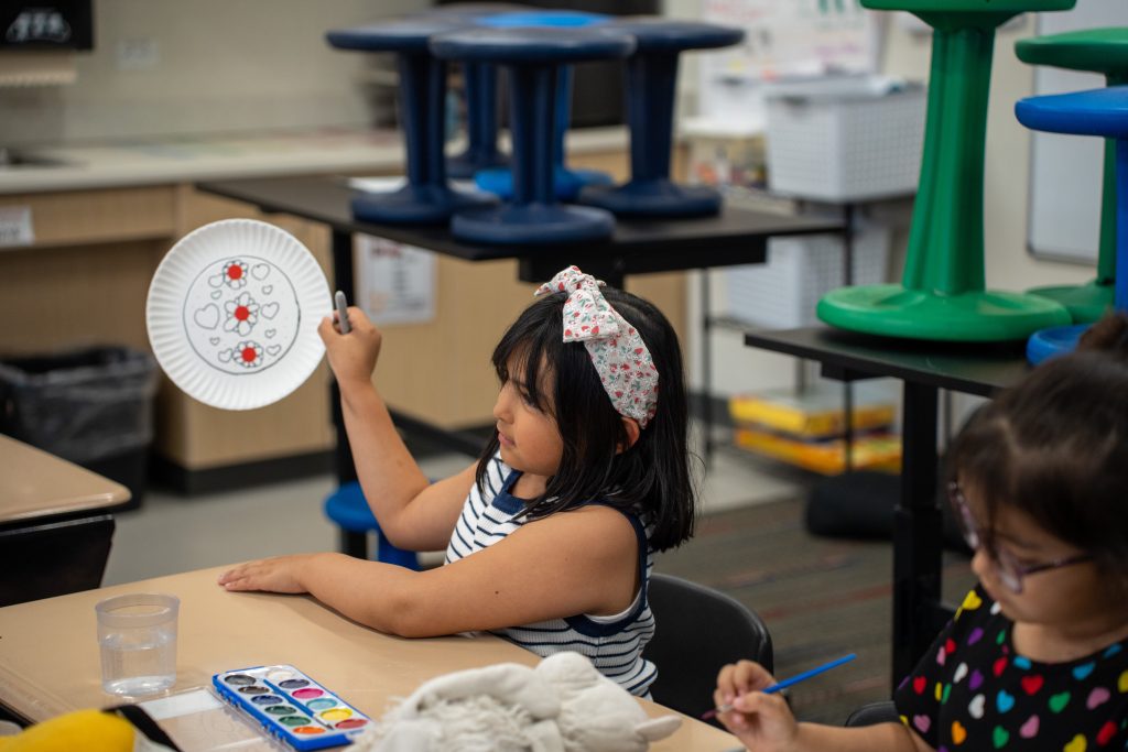 A girl holds up a decorated paper plate while another girl paints at a classroom table, with art supplies nearby.