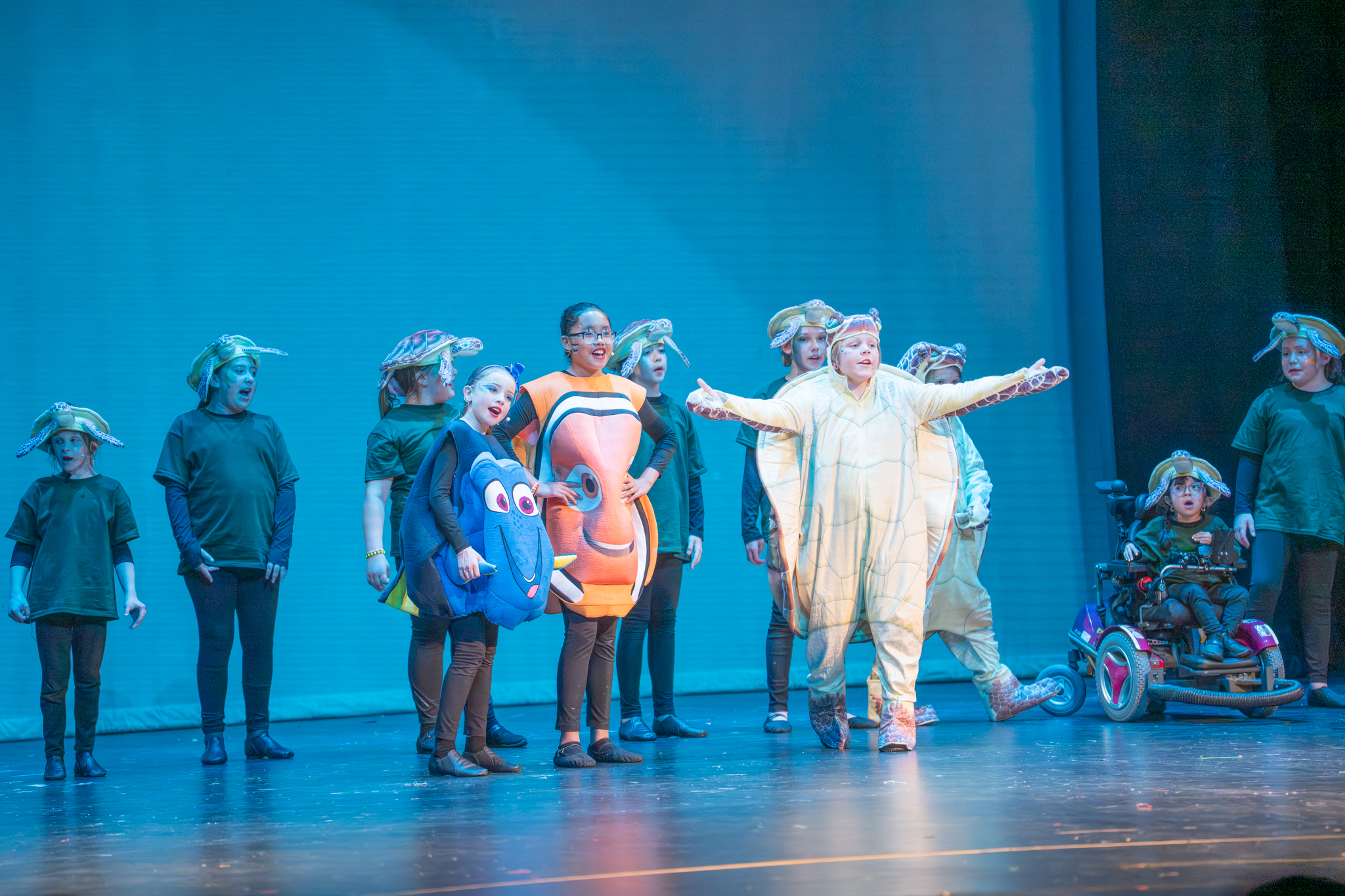 A group of children in costumes perform on stage, depicting sea creatures against a blue backdrop.