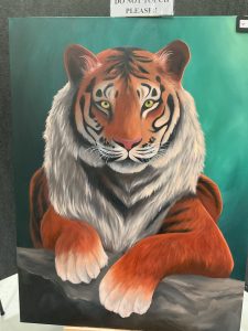 A realistic painting of a tiger with striking orange fur and intense green eyes, resting on a log against a teal background.