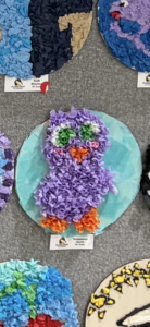 A colorful craft owl made of tissue paper, featuring purple body, green eyes, and orange beak, displayed on a wall.