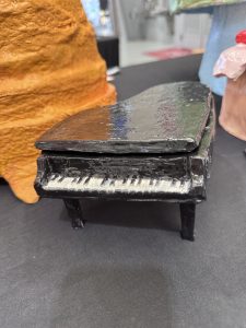 A small black grand piano model with white keys, displayed on a black surface with colorful sculptures in the background.