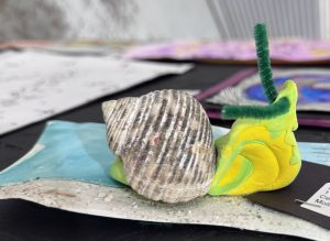 A colorful snail model made from clay and a shell, placed on a textured blue and sandy background.