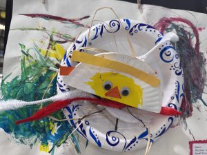 A craft project featuring a yellow chick made from paper plates and pipe cleaners, set against colorful painted background.