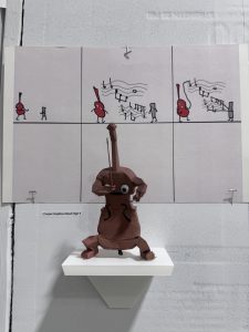 A clay figure playing a cello is displayed below a four-panel comic with musical themes.