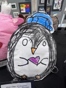 A handmade penguin drawing with a heart, displayed among other art pieces at an exhibition.