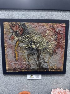 Textured artwork featuring warm colors and abstract shapes, displayed on a gray background with a nameplate below.