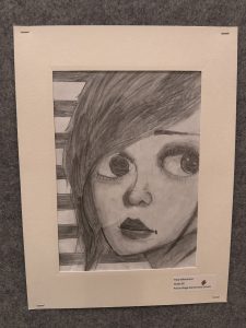 A pencil drawing of a girl with large eyes and dark hair, framed and displayed at an art exhibit.