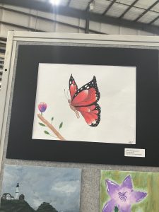 Un colorido dibujo de una mariposa con alas rojas y negras posada en una rama con una flor, enmarcado para exponerlo.