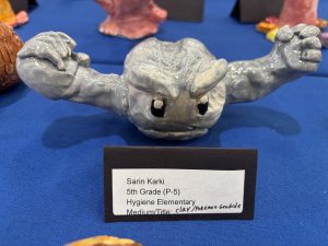 A clay sculpture of a gray Pokémon character with arms raised, displayed on a blue tablecloth at an art exhibit.