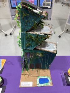 A three-tiered model with green paint, plants, and a blue pond, displayed on a purple table at an exhibit.