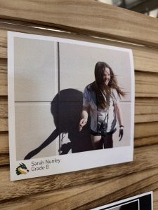 A smiling girl with long hair stands against a wall, holding a camera, with her shadow visible on the surface.