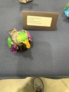 A colorful clay bird sculpture by a second grader, displayed on a table with an identification card.