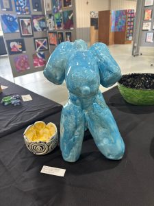 A blue ceramic sculpture stands next to a yellow and white bowl on a black table, with artwork displayed in the background.