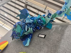 A colorful paper sculpture of a whale made from blue and green origami pieces, displayed on a gray backdrop.