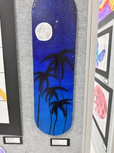 Una tabla de monopatín con un cielo nocturno en el que se ven una luna grande y siluetas de palmeras sobre un fondo azul.