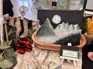 A handmade skeleton figure stands beside a mountain painting under a full moon, with a nameplate identifying the artist.