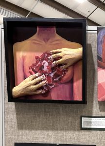 A painted figure with no head holds a textured, red heart, using hands with painted nails, set in a shadowed frame.