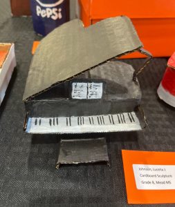A cardboard piano sculpture with a black body and keys, displayed on a table with an orange label.