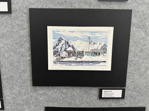 Una colorida ilustración de montañas nevadas y edificios de madera junto al agua, expuesta en una galería.