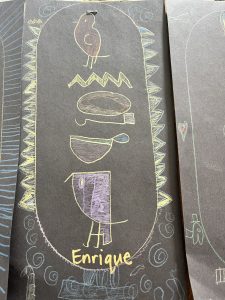 Child's artwork on dark paper featuring colorful drawings of a bird, bowls, and the name "Enrique" at the bottom.
