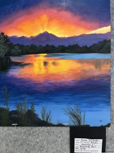 A vibrant sunset over mountains reflecting in a calm lake, with silhouettes of trees and grass in the foreground.