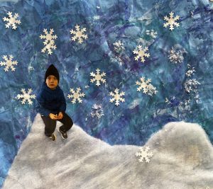 A child in a blue jacket sits on a snowy surface, surrounded by paper snowflakes against a blue background.