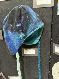 A colorful fleece hat with a galaxy pattern, featuring braided ties in shades of blue and green.