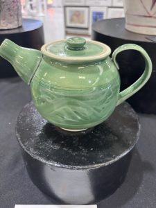 Green ceramic teapot with a lid, featuring a textured design, displayed on a black pedestal.