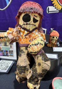 A handmade doll with patchwork clothing, featuring a sad face and a smaller doll beside it, displayed at an exhibit.