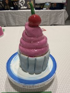 A colorful, handmade cupcake sculpture with pink frosting and a red cherry on top, displayed on a decorative plate.