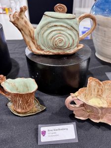 A snail-shaped teapot in green and brown, accompanied by two smaller ceramic cups, displayed on a black pedestal.