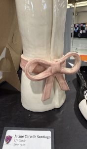 Ceramic vase with a glossy white finish and a pink bow tied around its center, displayed at an art exhibition.