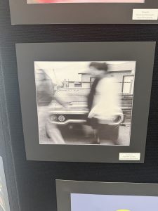 Black and white photograph of a car with blurred figures walking past it, displayed in a gallery setting.