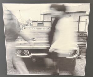 A blurred black-and-white photo of a classic car with two people walking past it.