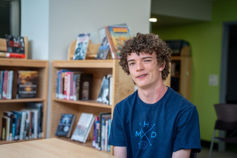 A young person with curly hair smiles while sitting at a table in a library, surrounded by bookshelves.