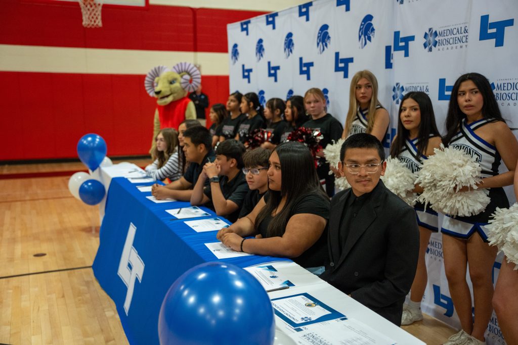 Students sit at a table with documents, while cheerleaders and a mascot stand behind them in a gym setting.