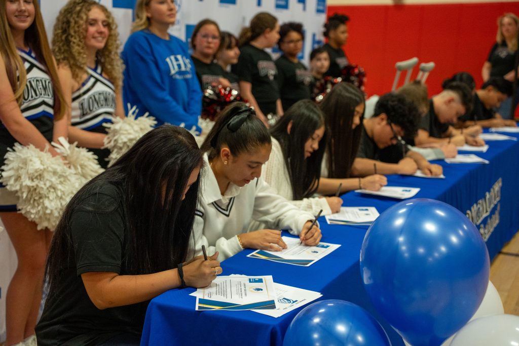 Students are signing documents at a table, with cheerleaders and supporters in the background. Blue balloons are visible.