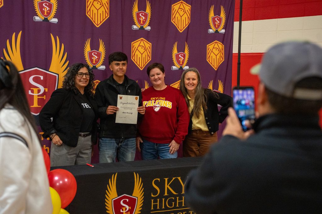Four people pose for a photo at a celebration event, holding a certificate, with a school backdrop and balloons.
