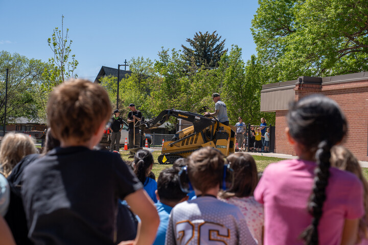 Children watch a demonstration with heavy machinery in a schoolyard on a sunny day. Trees and a building are in the background.