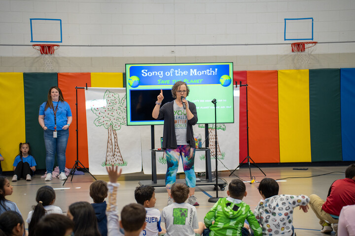 A teacher leads a music session about environmental awareness in a colorful gym, with students engaged on the floor.