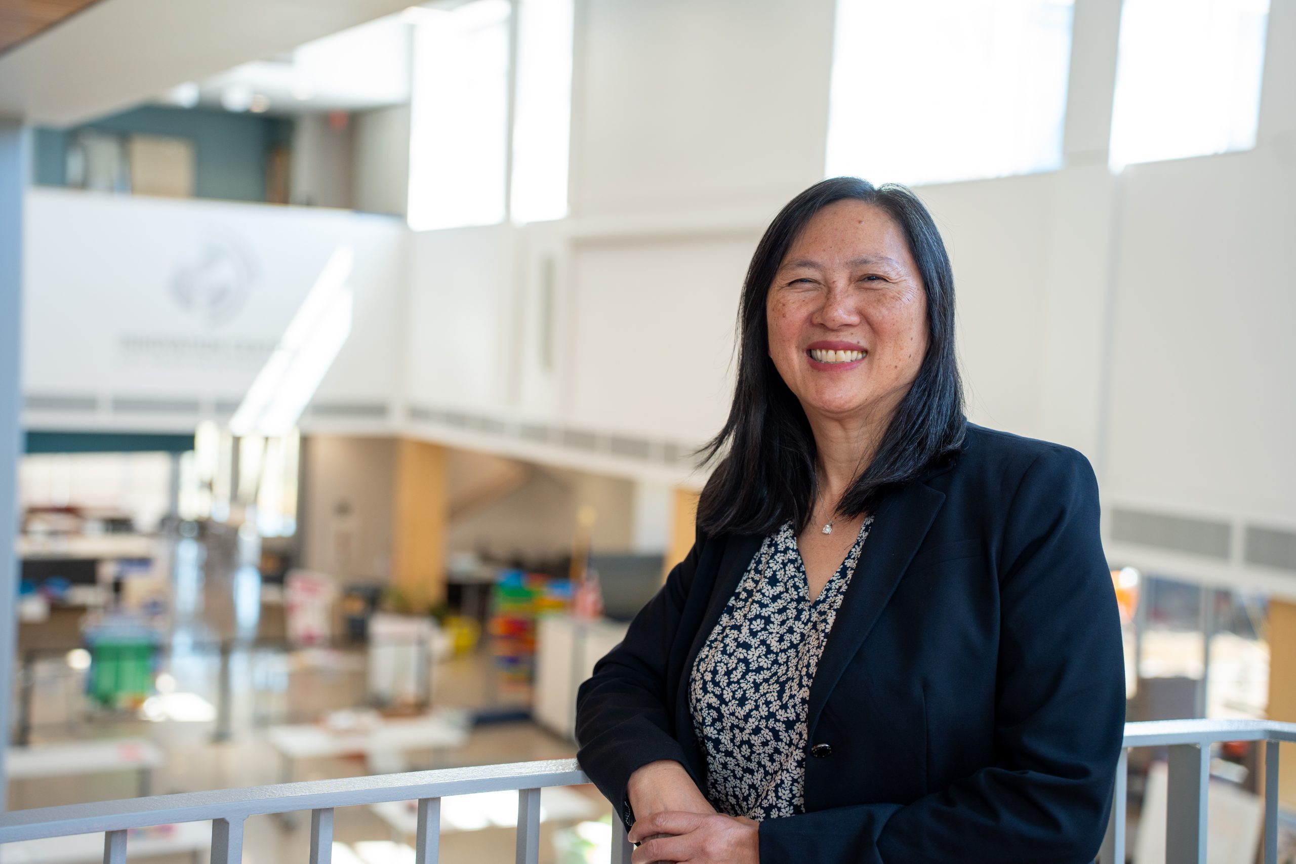 A woman in a black blazer smiles while standing by a railing in a bright, open indoor space with colorful objects in the background.