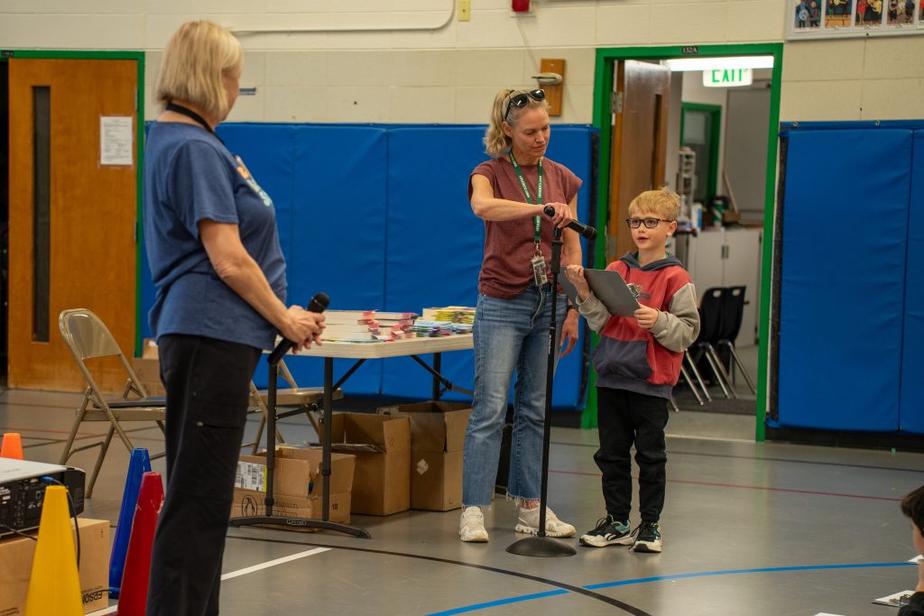 A student speaks at a microphone while his teacher stands nearby, holding a clipboard, in a school gym setting.