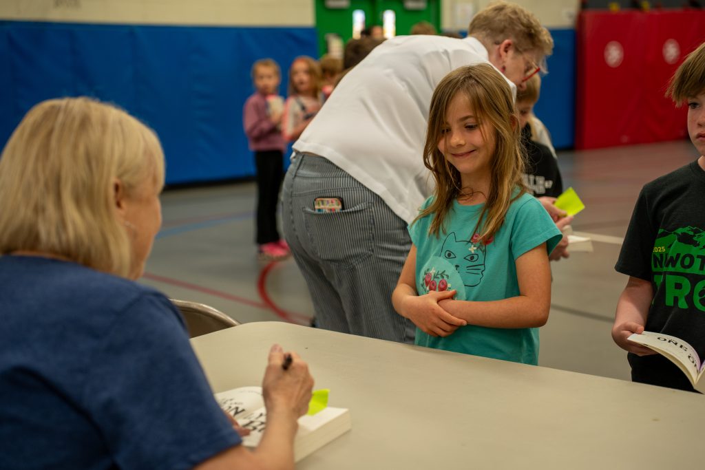 A young girl smiles while waiting in line at the book signing event, with Katherine Applegate signing books at a table.