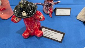 A red clay alien sculpture with big eyes and a black hat, displayed on a blue table with name tags.