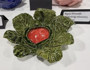 A ceramic dish shaped like a green leaf, featuring a red center, displayed at an art show.